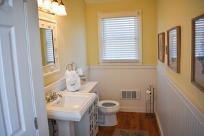 Une salle de bain avec un lavabo, des toilettes et un meuble-lavabo, tous de couleur blanche. On y trouve une fenêtre avec des stores, un miroir et des tableaux encadrés accrochés au mur.
