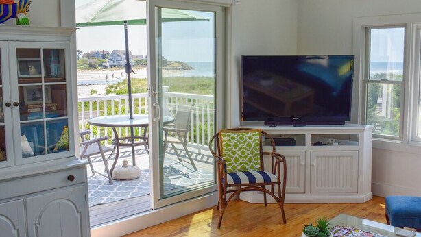 A living room with a TV, a dining table, and a chair. There's a glass door leading to a balcony with a view of the beach. - Three bedroom sleeping 6 just 150 steps to South Village Beach (West Dennis)