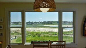 A dining area with a table and chairs, large windows with a view of a beach and water, and a woven light fixture above. - Three bedroom sleeping 6 just 150 steps to South Village Beach (West Dennis)