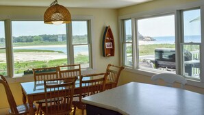 A dining area with a table and chairs, large windows with a view of the beach and ocean, and a hanging light fixture. - Three bedroom sleeping 6 just 150 steps to South Village Beach (West Dennis)