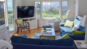 A living room with a large TV, white furniture, and a view of greenery outside. - Three bedroom sleeping 6 just 150 steps to South Village Beach (West Dennis)