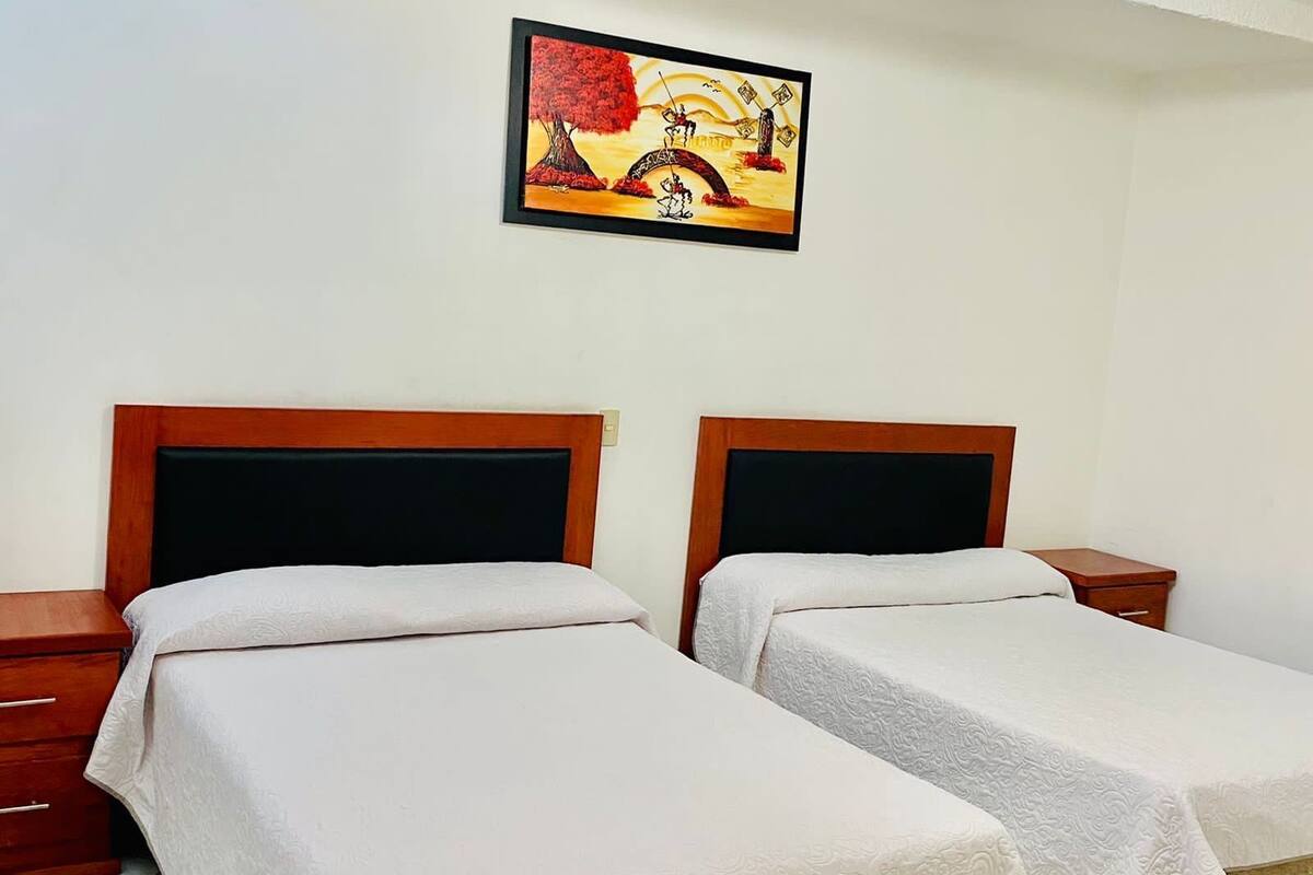 A hotel room with two single beds, wooden headboards, white bedding, a nightstand, and a framed painting on the wall.