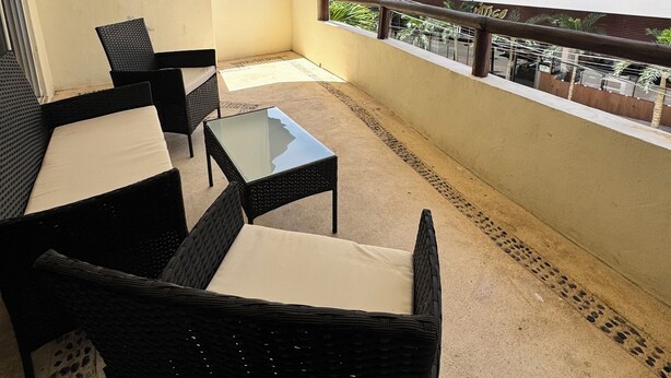 A balcony with wicker furniture and a glass table, overlooking a building with multiple balconies and windows.