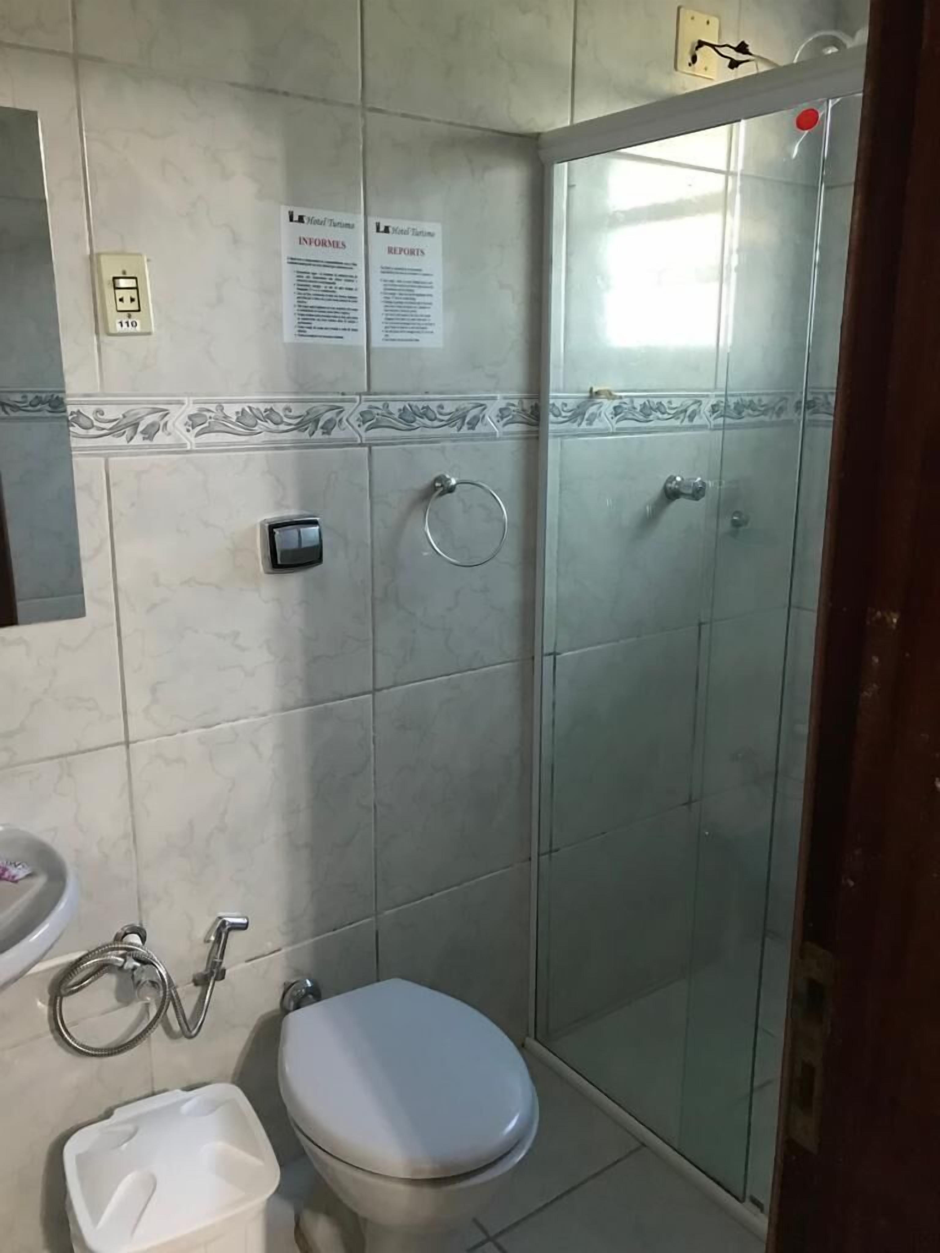Baño con inodoro, bidé y cabina de ducha. Hay señales informativas en la pared y un enchufe.
