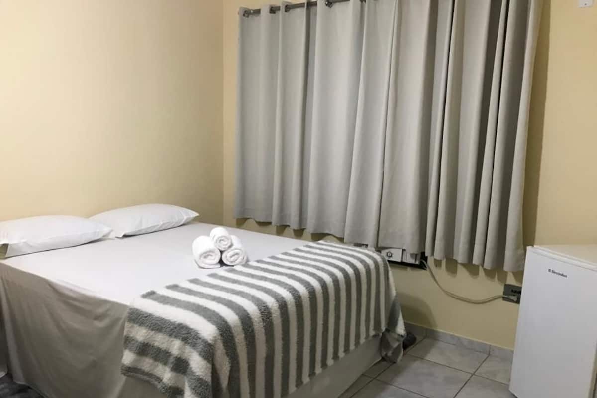 A hotel room with a bed, a striped blanket, rolled towels, a curtain, a radiator, and a refrigerator.