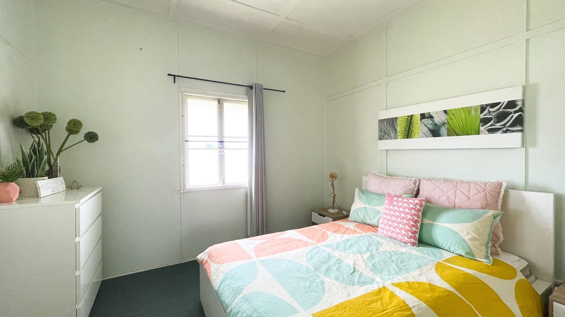 A bedroom with a bed, a bedside table, a nightstand, a window with curtains, a wall-mounted artwork, and a dresser with plants.