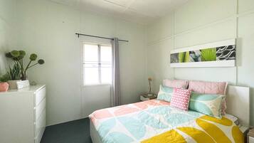A bedroom with a bed, a bedside table, a nightstand, a window with curtains, a wall-mounted artwork, and a dresser with plants.