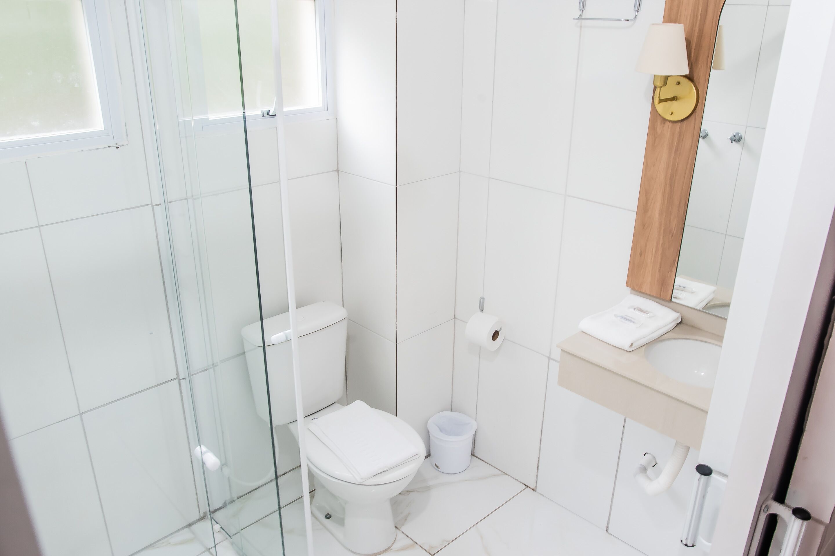 A modern bathroom with a glass shower enclosure, a white toilet, a sink with a wooden vanity, and a mirror with a clock.