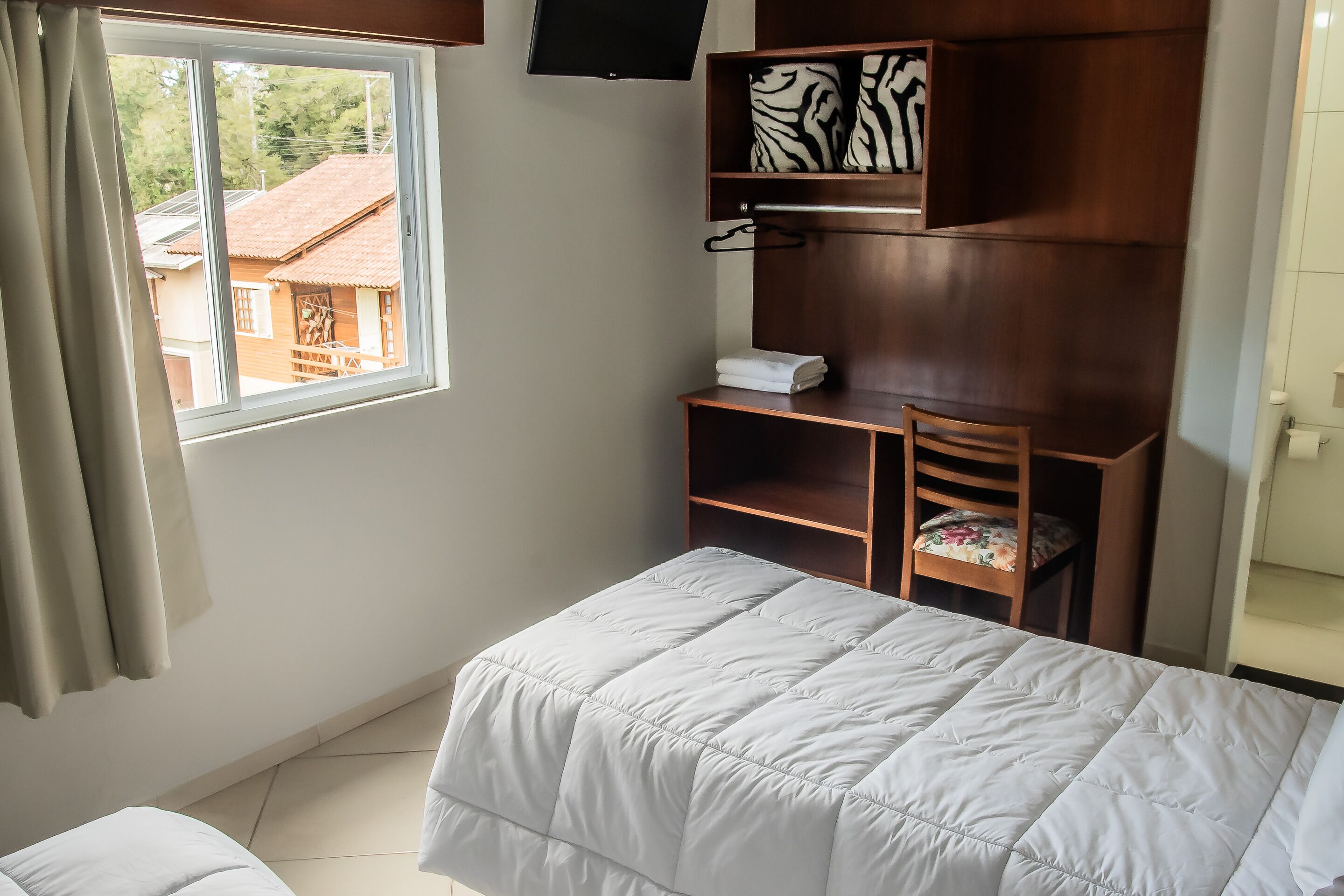 A bedroom with a bed, a desk, and a chair. There is a window with curtains, a shelf with a zebra-patterned pillow, and a television on the wall.