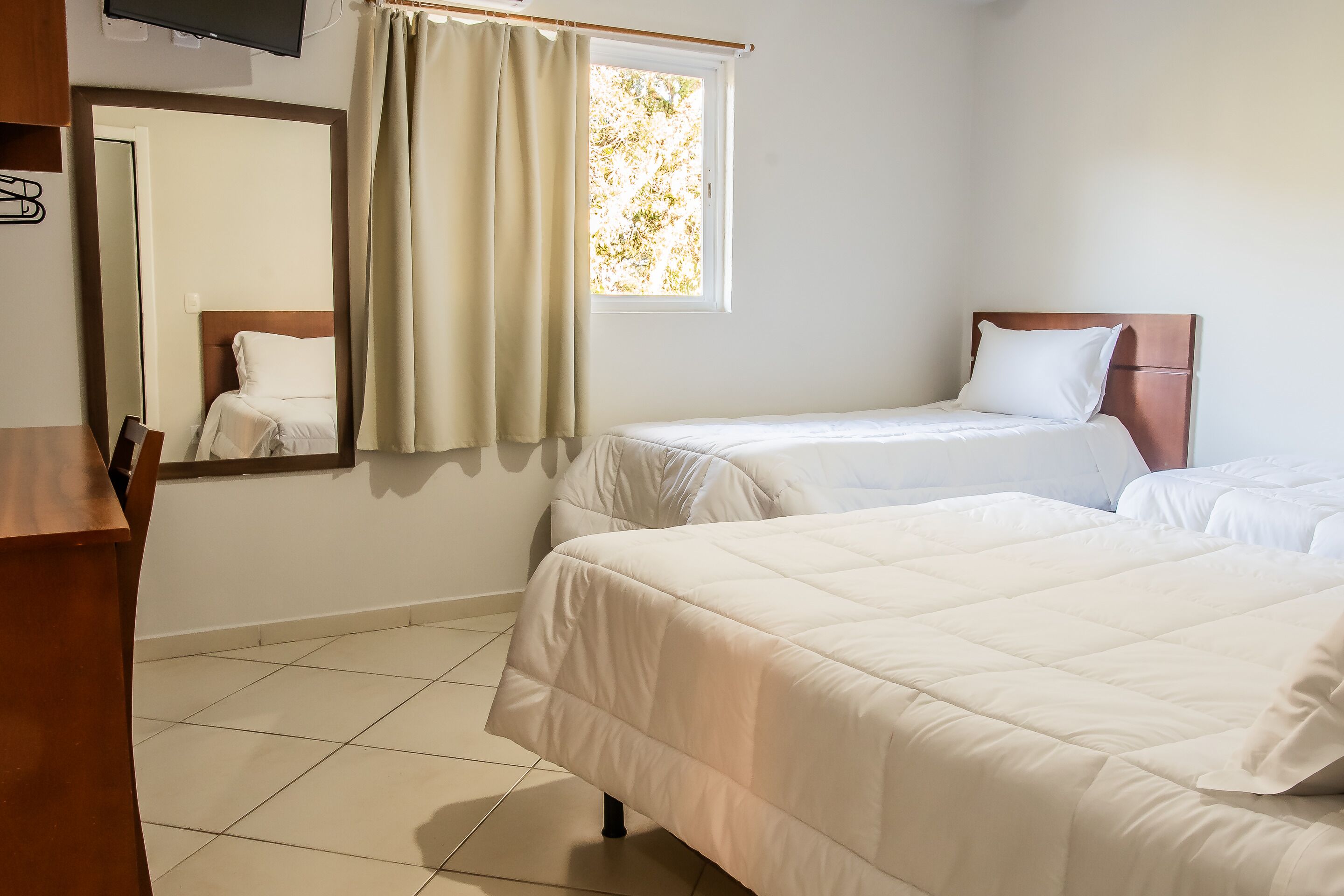 Habitación de hotel con dos camas, cabecero de madera, ventana con cortinas, televisor de pantalla plana montado en la pared y suelo de baldosas.