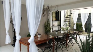 A dining area with a wooden table and chairs, surrounded by white curtains. There are potted plants and a shelf with decorative items. - Mi Ca-Sa En Mérida (Mérida)