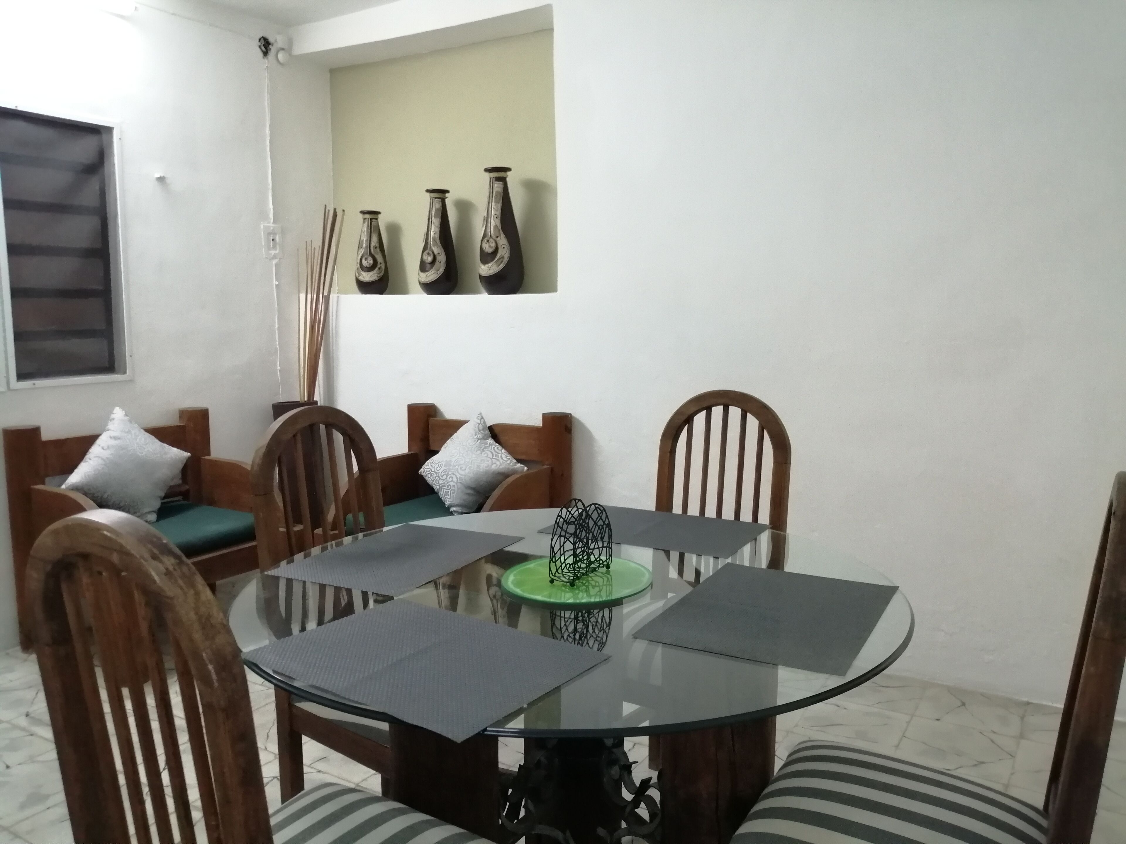 Un coin repas avec une table ronde en verre, des chaises en bois et une nappe verte. Trois vases sont accrochés au mur, au-dessus de la table.