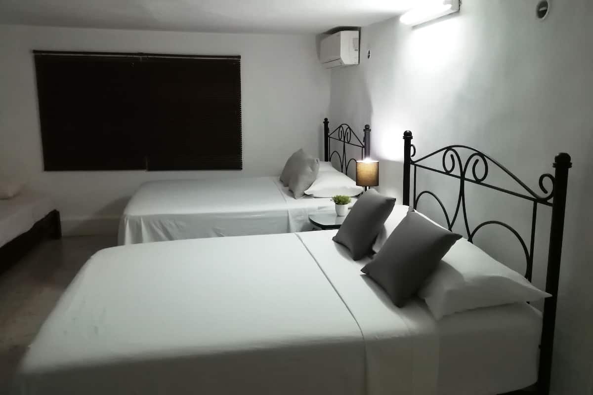 Une chambre avec deux lits simples, une table de chevet avec une lampe, une fenêtre avec des stores et un climatiseur fixé au mur.