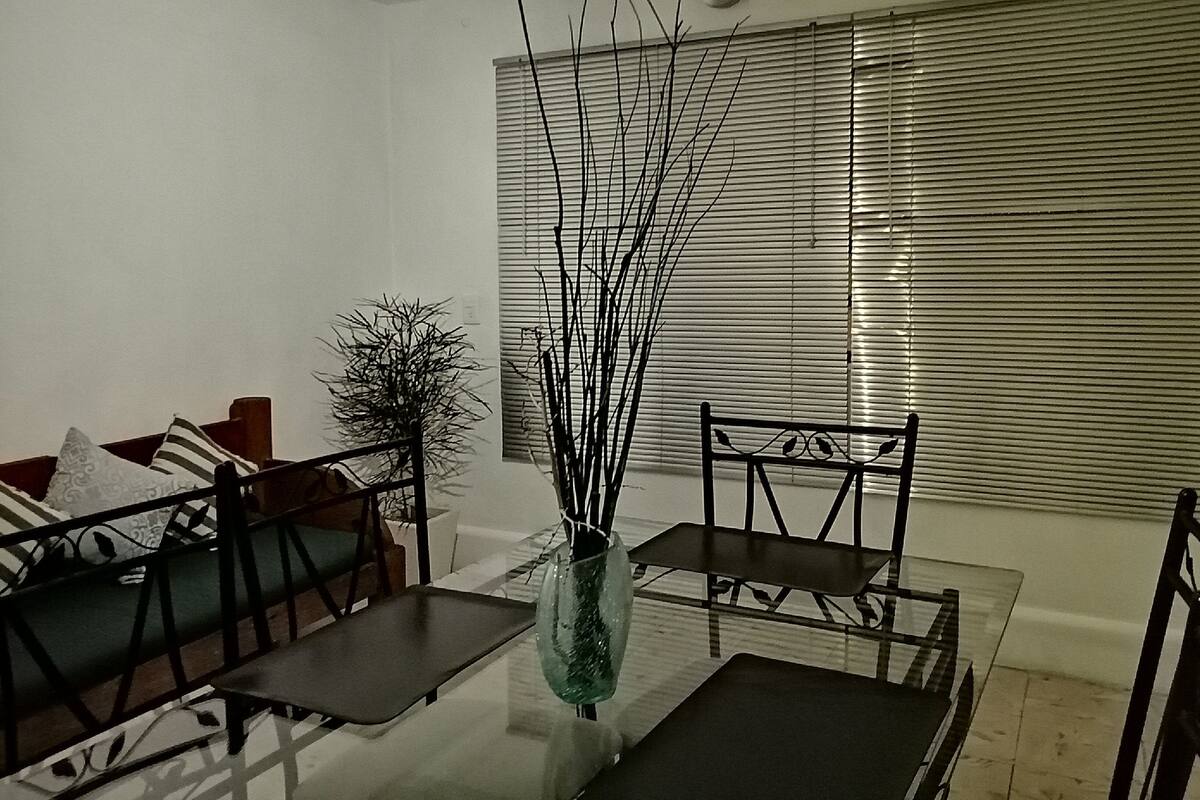 Un coin repas avec une table en verre, des chaises en métal et un vase décoré de branches. On aperçoit un canapé avec des coussins à motifs et une plante en pot à l'arrière-plan.