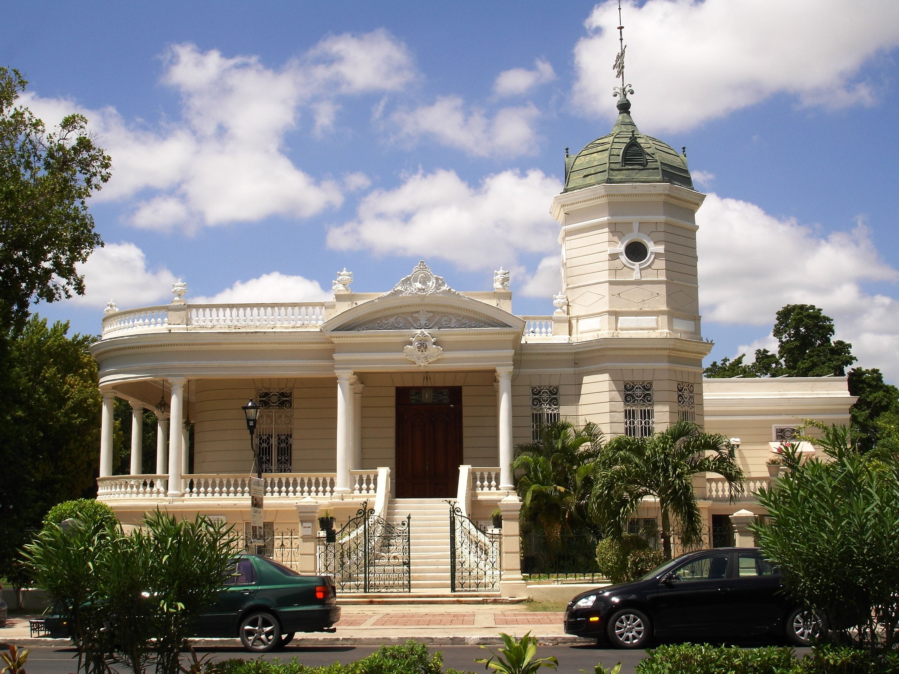 A grand building with a central dome and a tower, surrounded by lush greenery and parked cars.