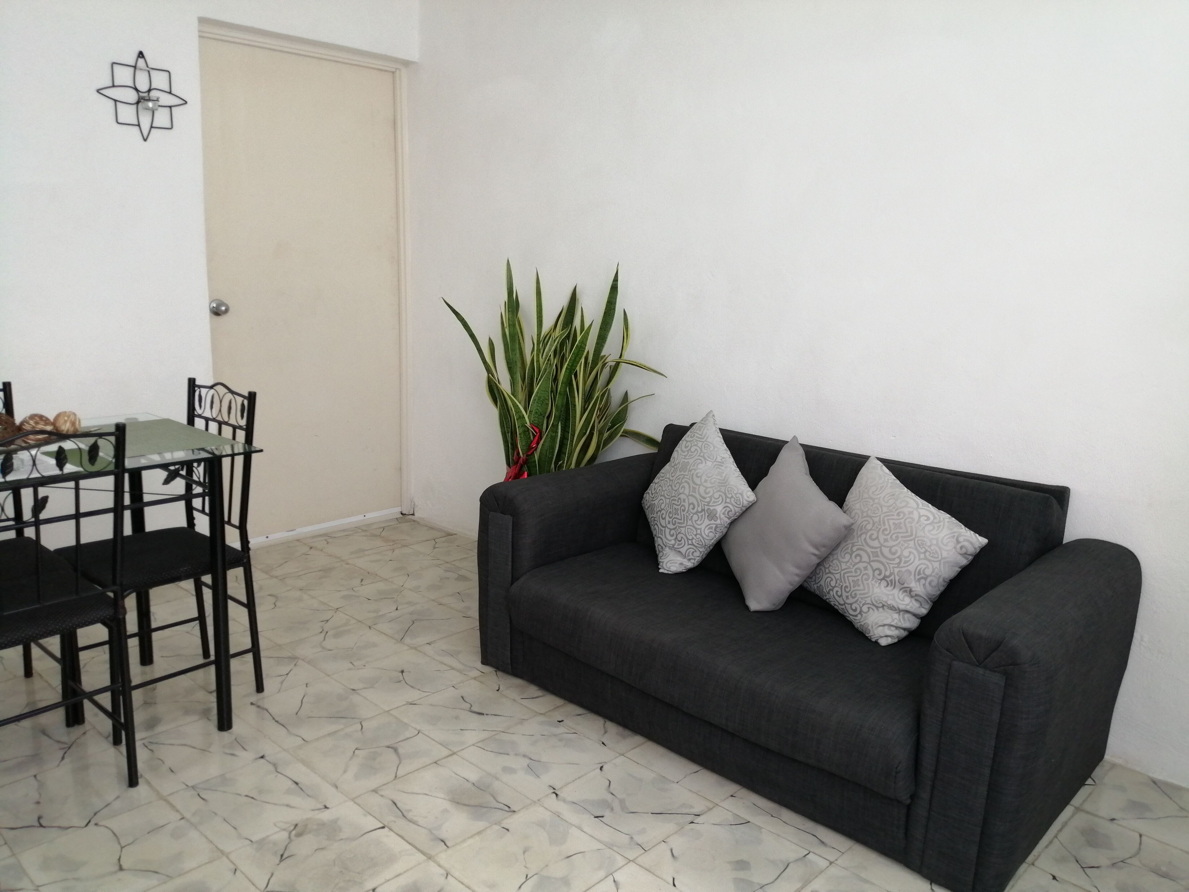 A living room with a black sofa, three grey pillows, a dining table with chairs, a potted plant, and a wall-mounted light fixture.