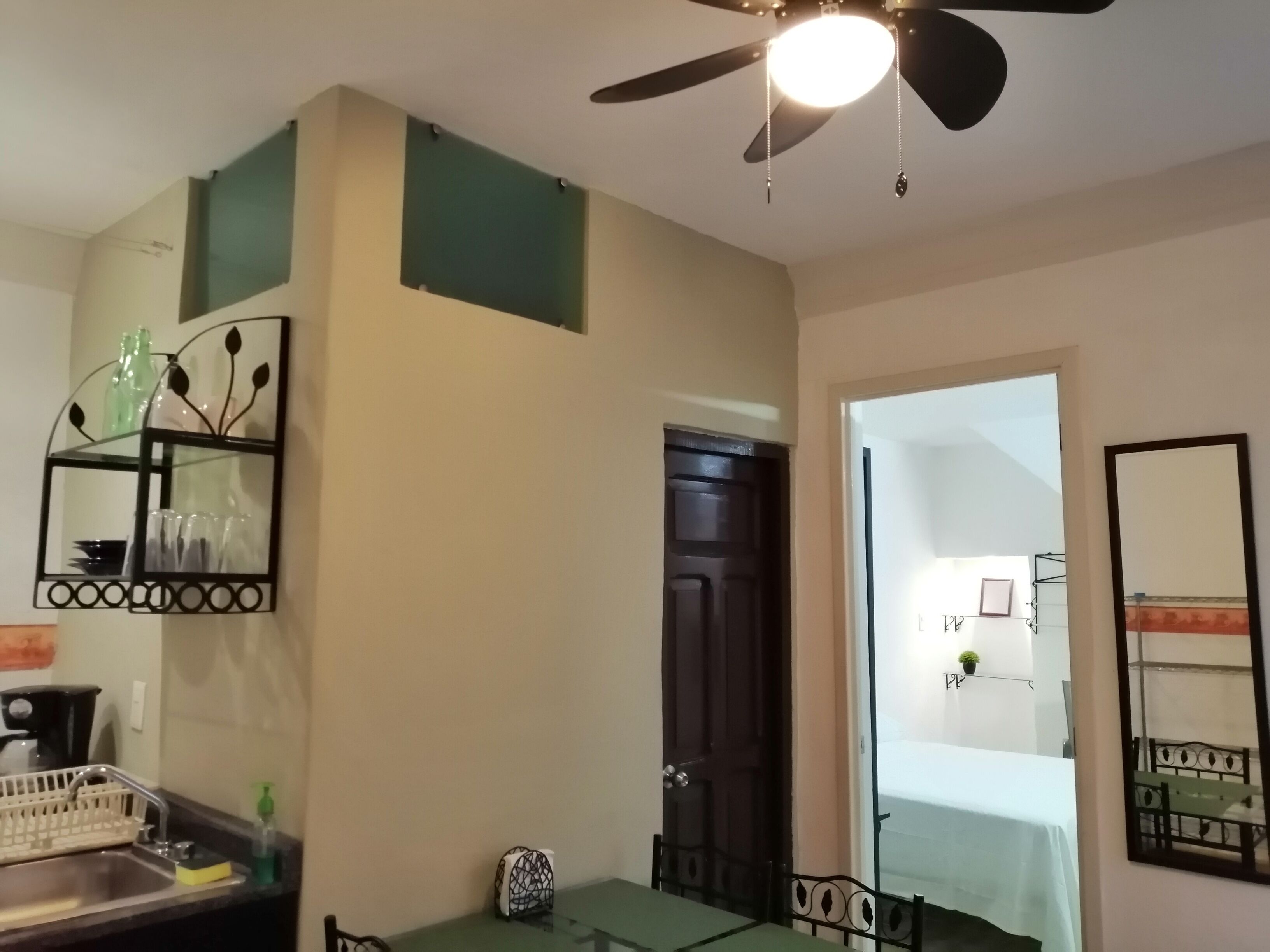 A room with a ceiling fan, a glass table, a metal chair, a sink with a dish rack, and a door leading to another room.