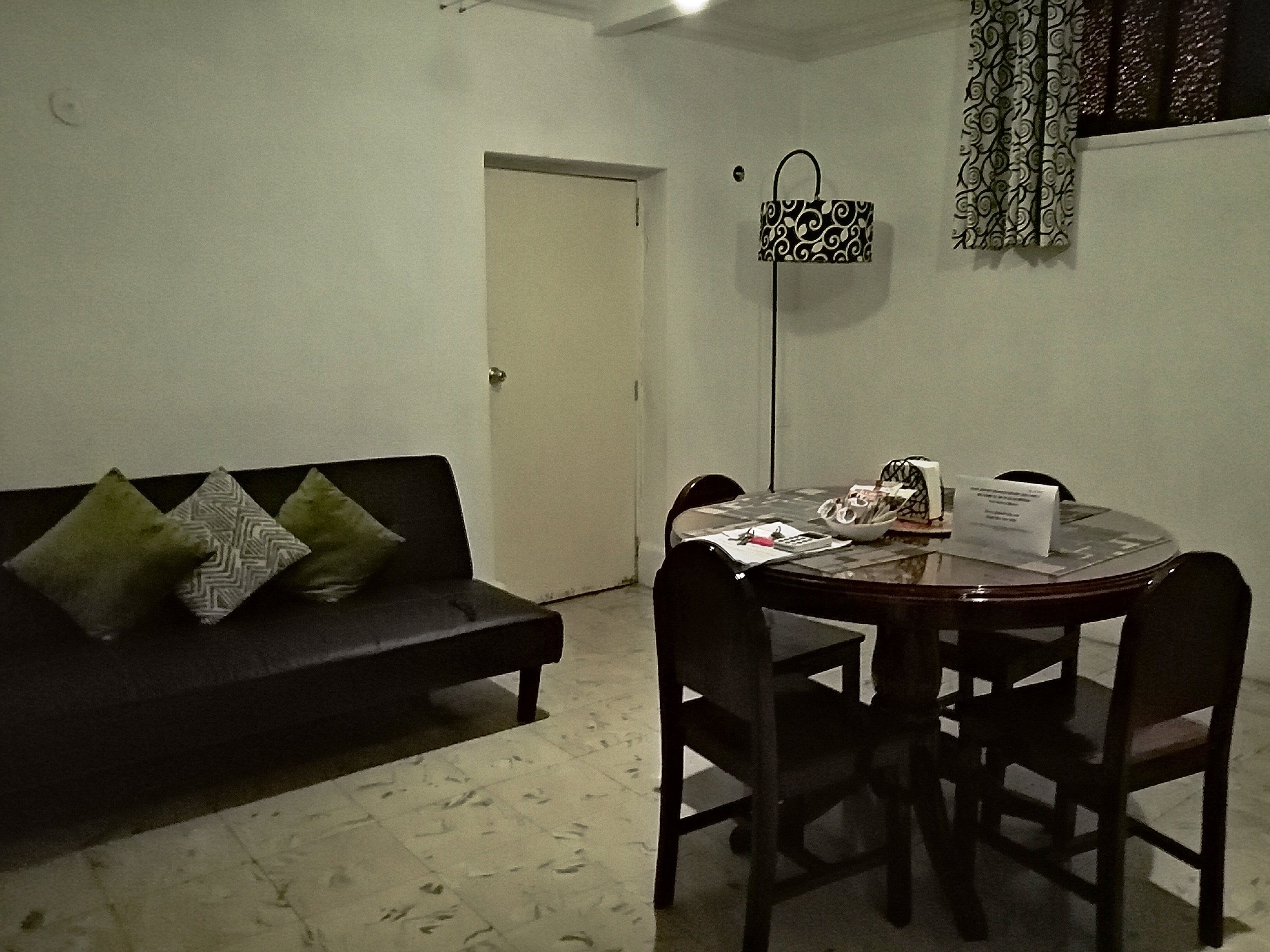 A living room with a black sofa, two patterned pillows, a dining table with chairs, a floor lamp, and a door.