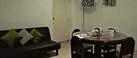 A living room with a black sofa, two patterned pillows, a dining table with chairs, a floor lamp, and a door.