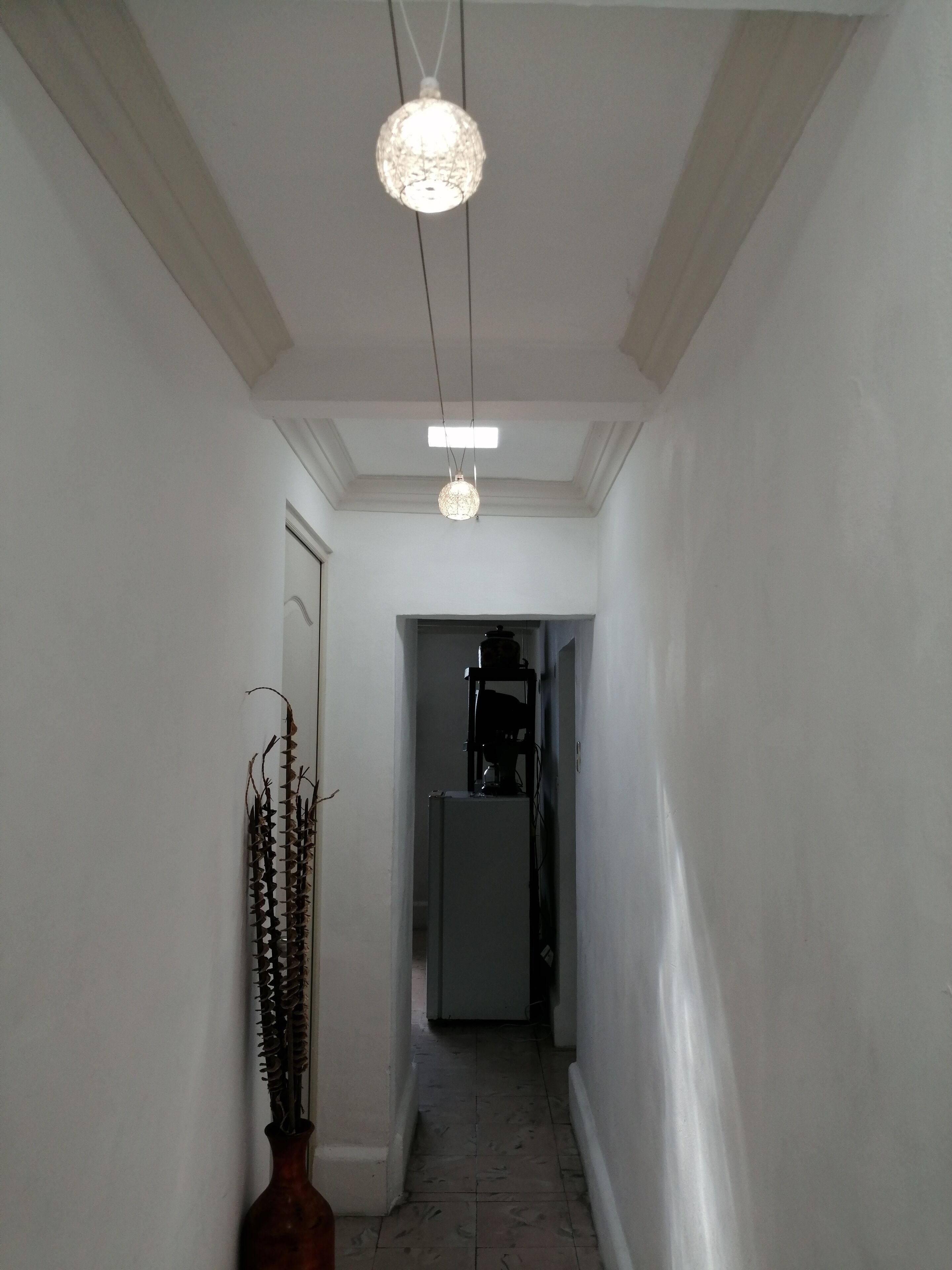 Ein schmaler Korridor mit weißen Wänden und Fliesenboden. Eine Hängelampe ist zu sehen, und eine hohe Vase mit Trockenpflanzen steht an der Wand.