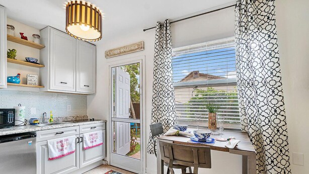 A kitchen with white cabinets, a microwave, a sink, and a dining table with chairs. There is a window with patterned curtains and a view of the outside. - Heart of Wellington  (Wellington)
