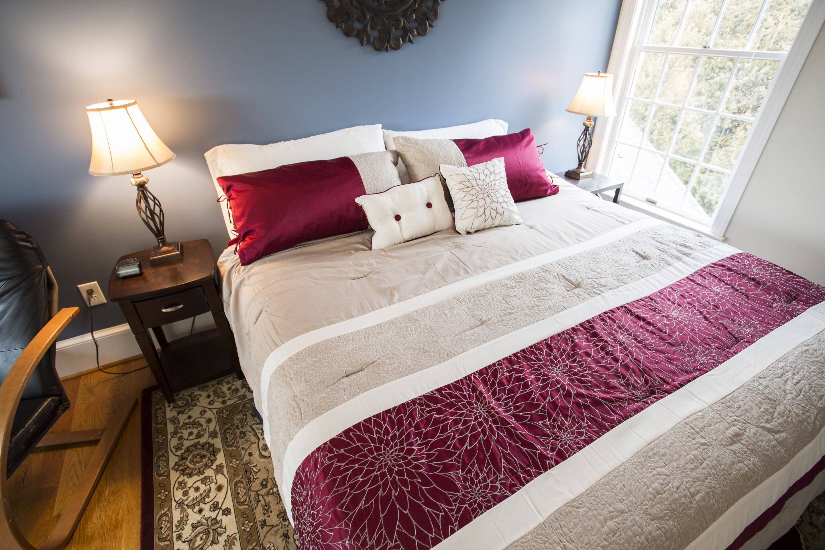 A neatly made bed with a patterned bedspread, two bedside tables with lamps, and a view of greenery through a window.