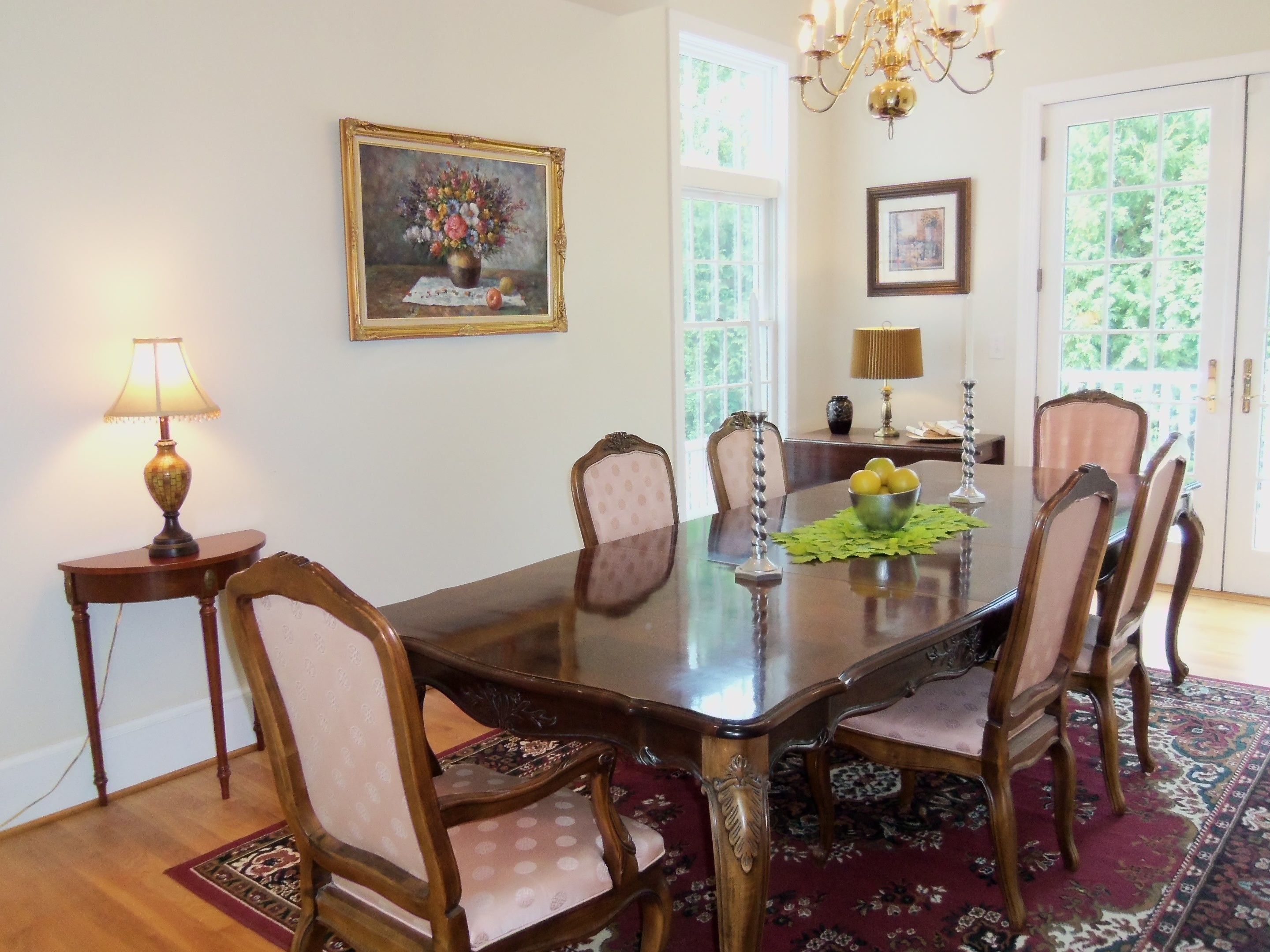 A dining room with a wooden table, six chairs, a chandelier, and a painting on the wall.