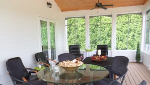 A covered outdoor dining area with a glass table surrounded by chairs, a candle centerpiece, and a view of greenery through large windows.