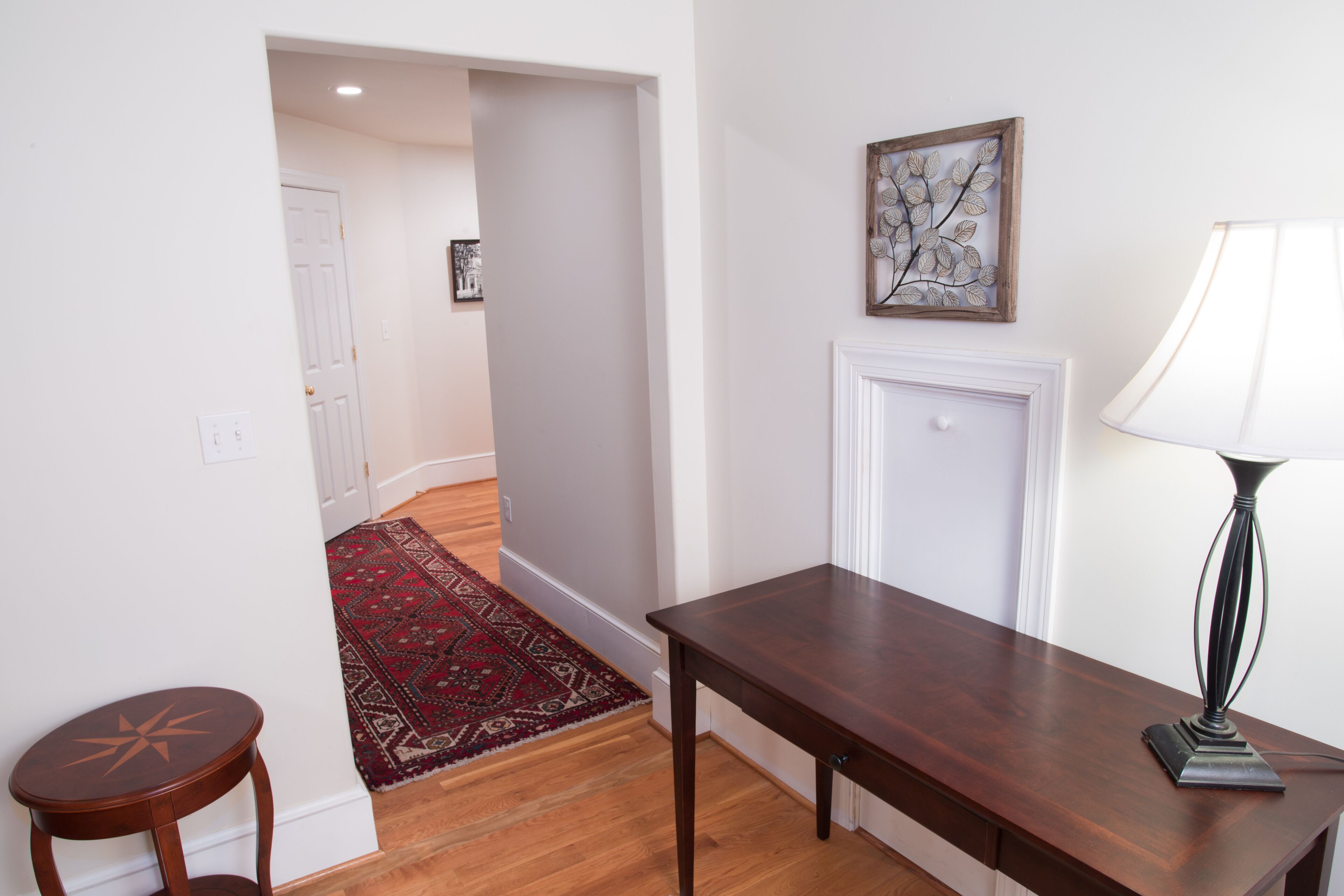 Una habitación con una mesa de madera, una lámpara, un tapete rojo y una puerta que da a otra estancia.