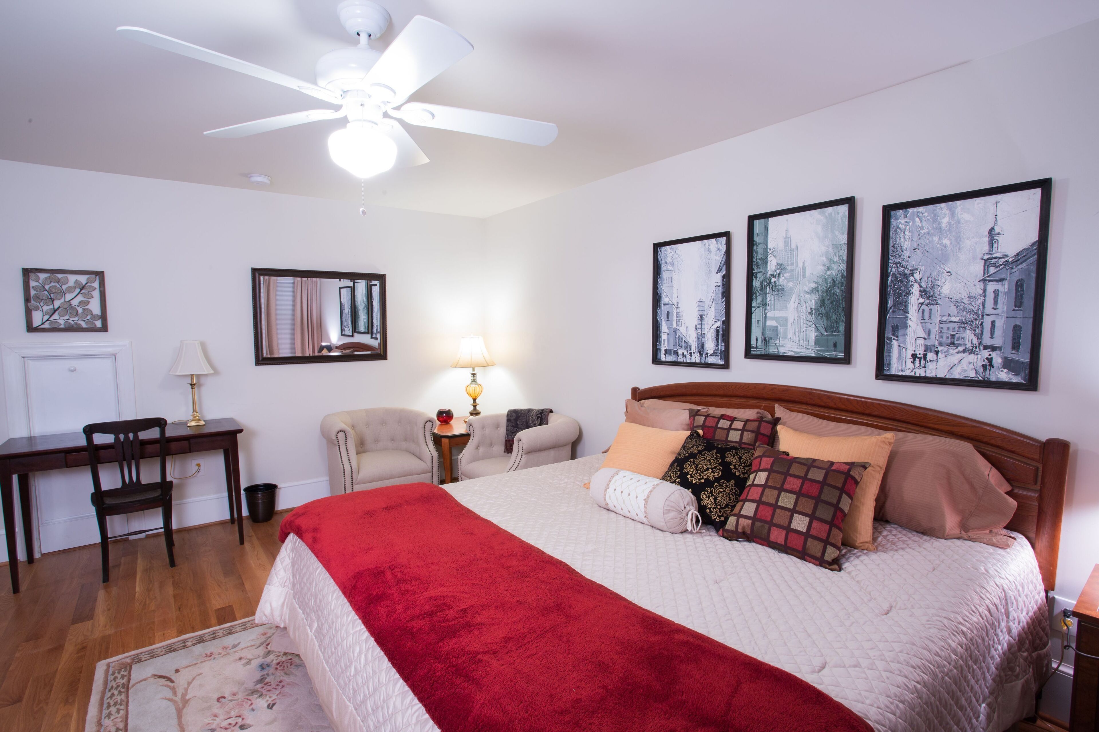 A neatly arranged bedroom with a bed, a desk, a chair, a lamp, and framed pictures on the wall.