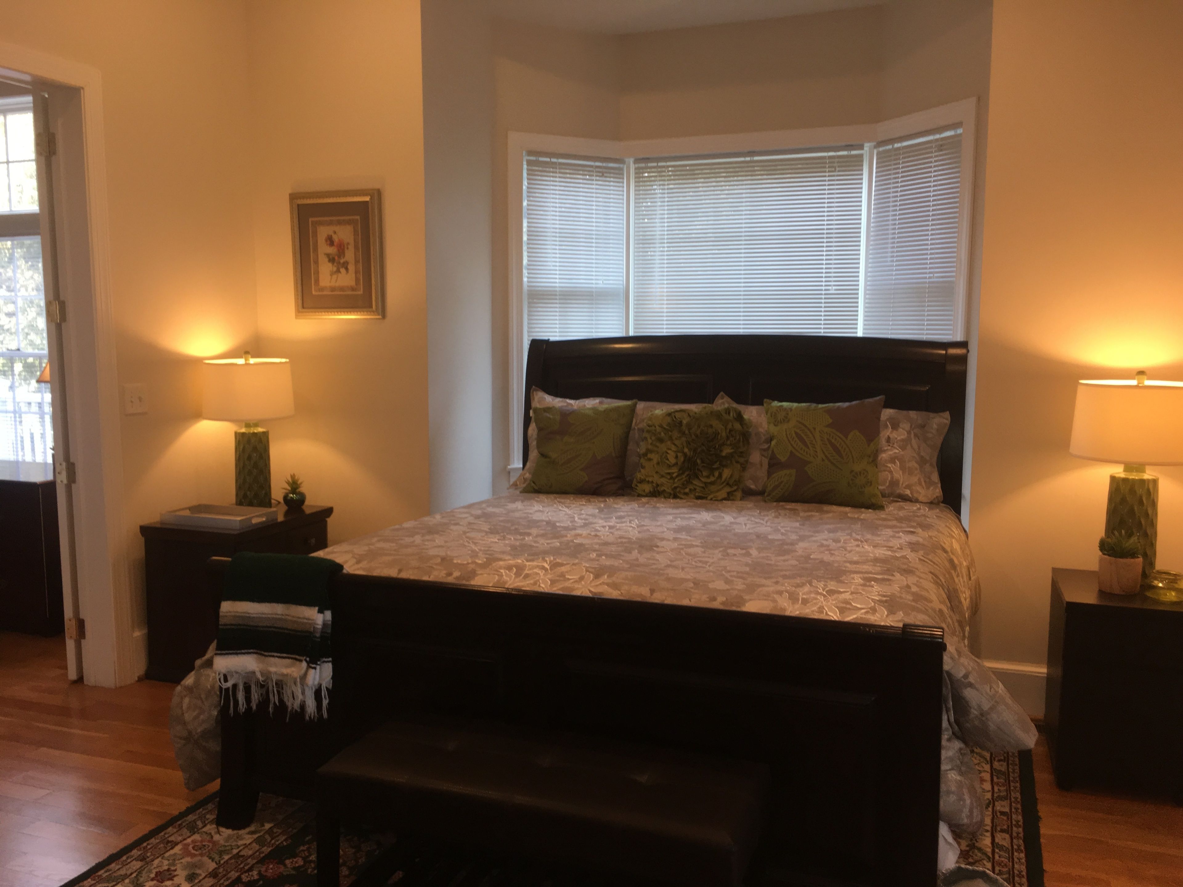 Une chambre à coucher avec un grand lit, deux tables de chevet, un banc, une fenêtre avec des stores et une porte.