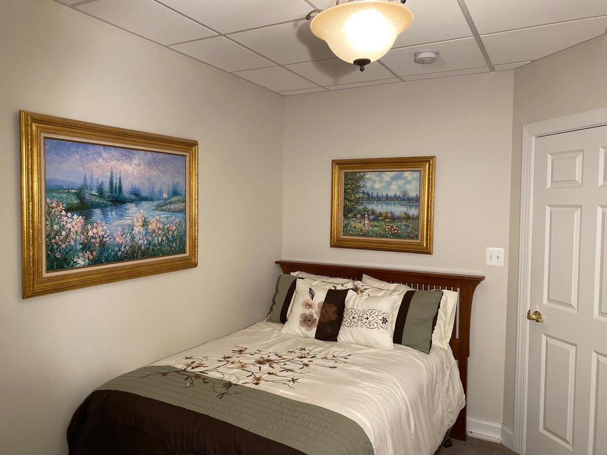 A bedroom with a bed, two framed paintings, a ceiling light, and a door.