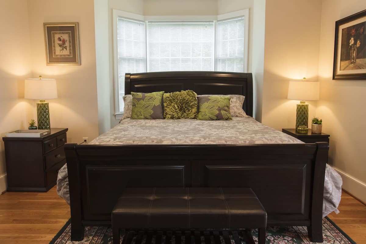 A bedroom with a dark wooden bed, green and white patterned bedding, two bedside lamps, a small plant on the nightstand, and a window with blinds.