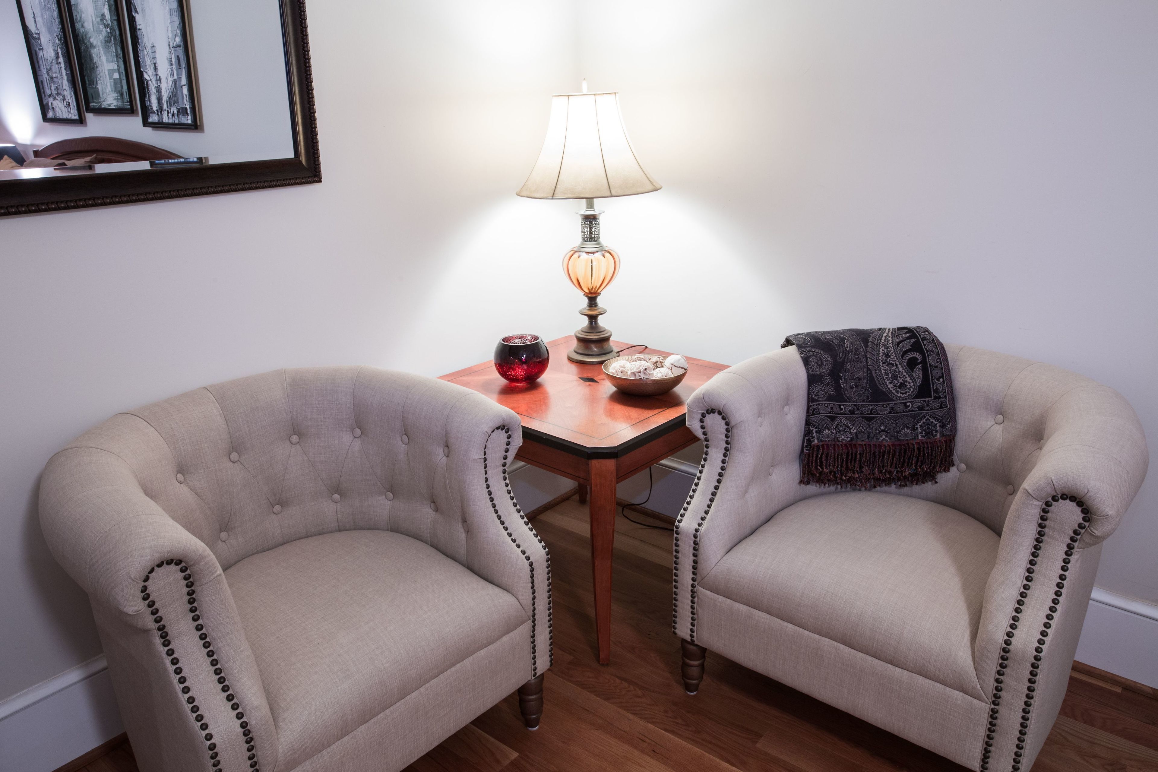 A room with two upholstered armchairs, a wooden table with a lamp, a bowl, and a red vase.