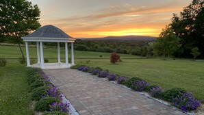 A gazebo with a brick pathway leading to it, surrounded by a grassy area with purple flowers.