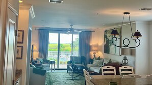 A spacious living room with a large window, a ceiling fan, and a dining area with a kitchen counter. - Great summer rates, Location, Views, Convenience...Golf/Pool/Fitness/Biking (Saint Simons Island)