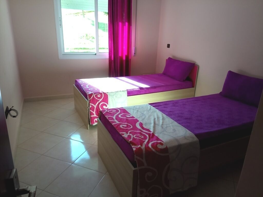 A room with two beds, each with a purple mattress and a patterned bedspread. There is a window with pink curtains, and the room has white tiled flooring.