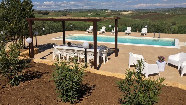 Ein Poolbereich mit Pergola, weißen Liegestühlen und einem Tisch mit Stühlen.