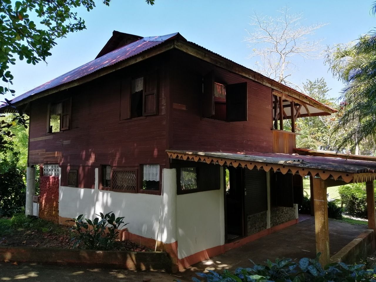 A two-story wooden house with a porch, surrounded by greenery.