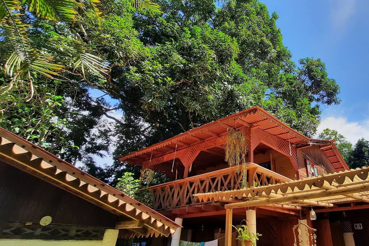 Une construction traditionnelle en bois, dotée d’un balcon, entourée d ’une végétation luxuriante et surplombant un ciel dégagé.