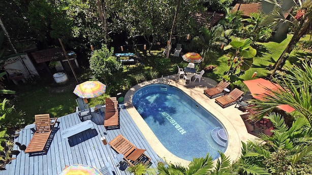 An outdoor swimming pool with lounge chairs and umbrellas, surrounded by lush greenery and a wooden deck. - Chalet Y Cabinas Hibiscus (Cahuita)