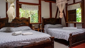 A room with two wooden beds, each with a floral bedspread and a rolled towel. There are lace curtains on the windows, and a mosquito net hanging above the beds. - Chalet Y Cabinas Hibiscus (Cahuita)