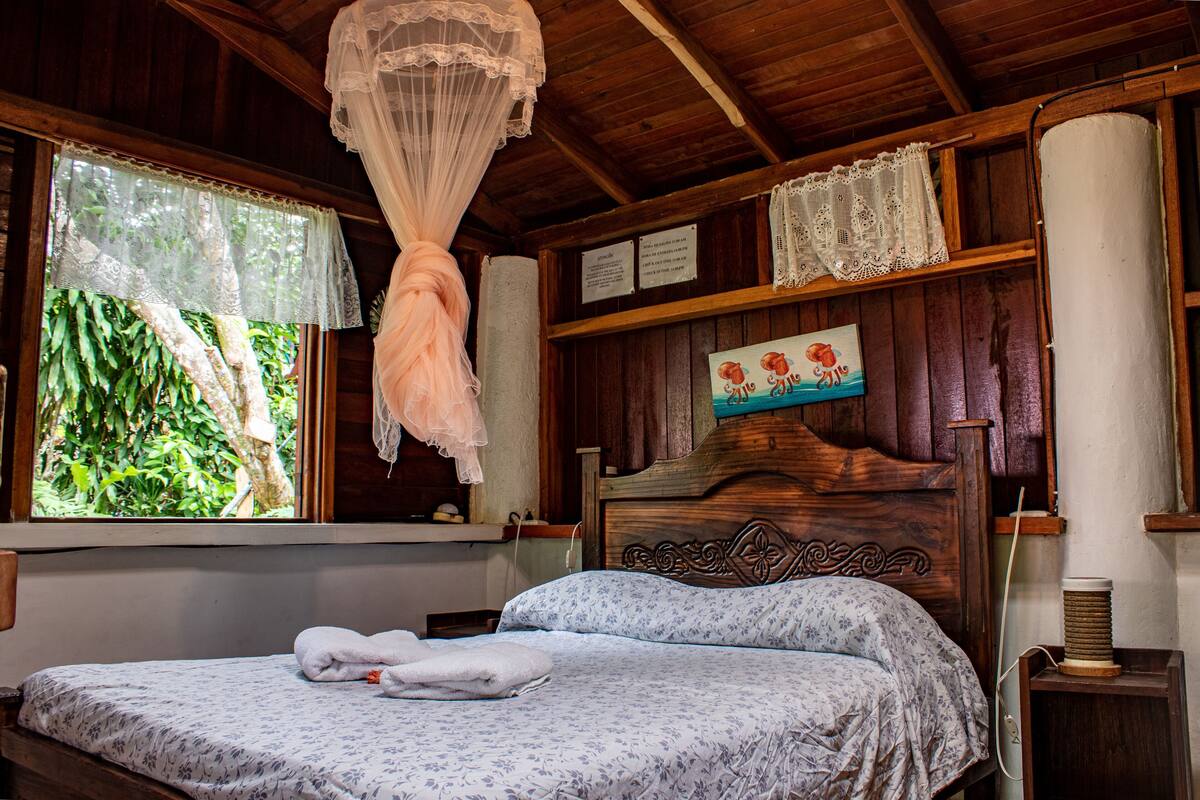 Une pièce en bois avec un lit, une fenêtre ornée de rideaux en dentelle et un mur sur lequel est accroché un tableau encadré.