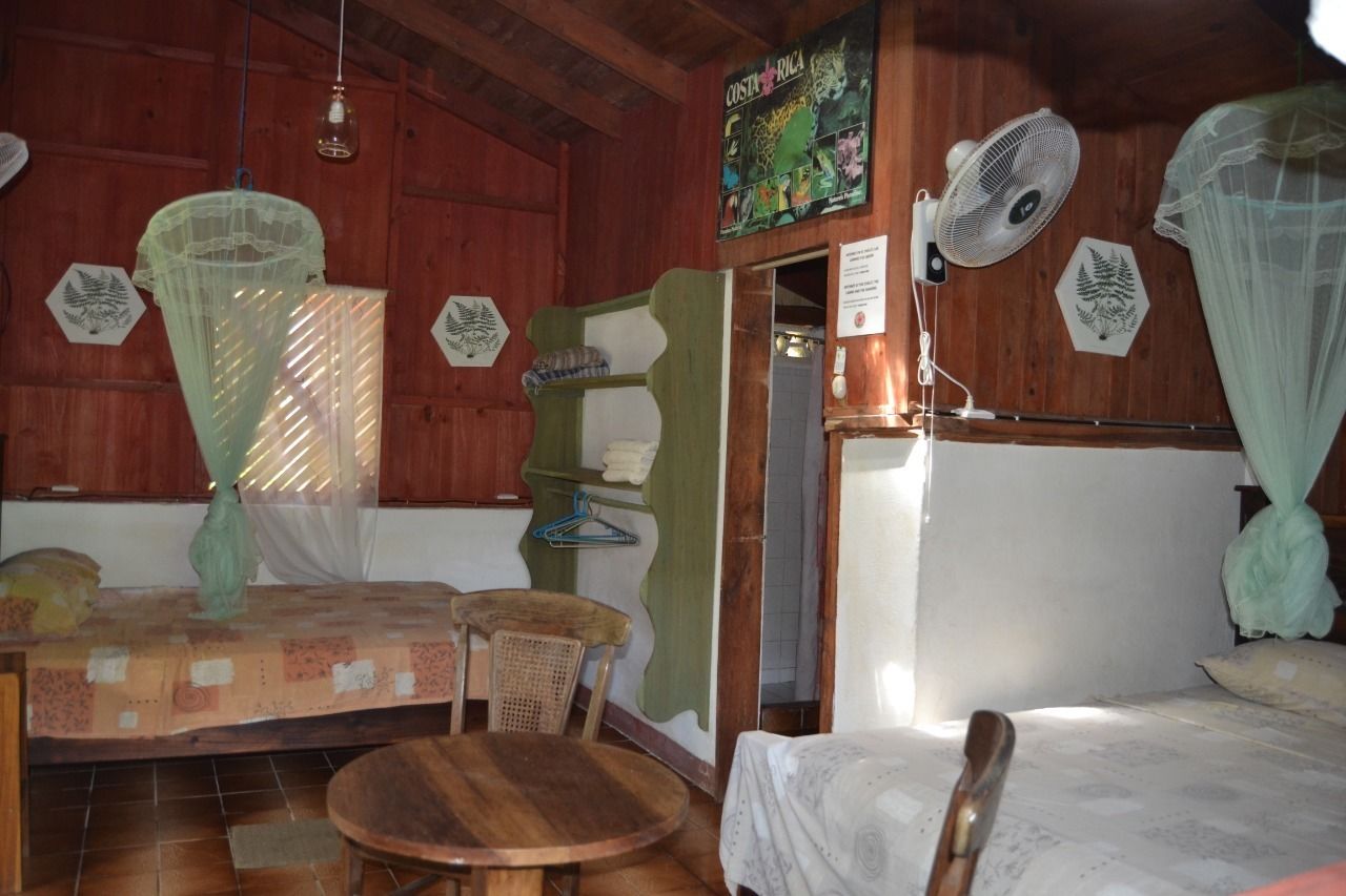 A room with wooden walls, a bed with a mosquito net, a table, a chair, and a wall-mounted fan.