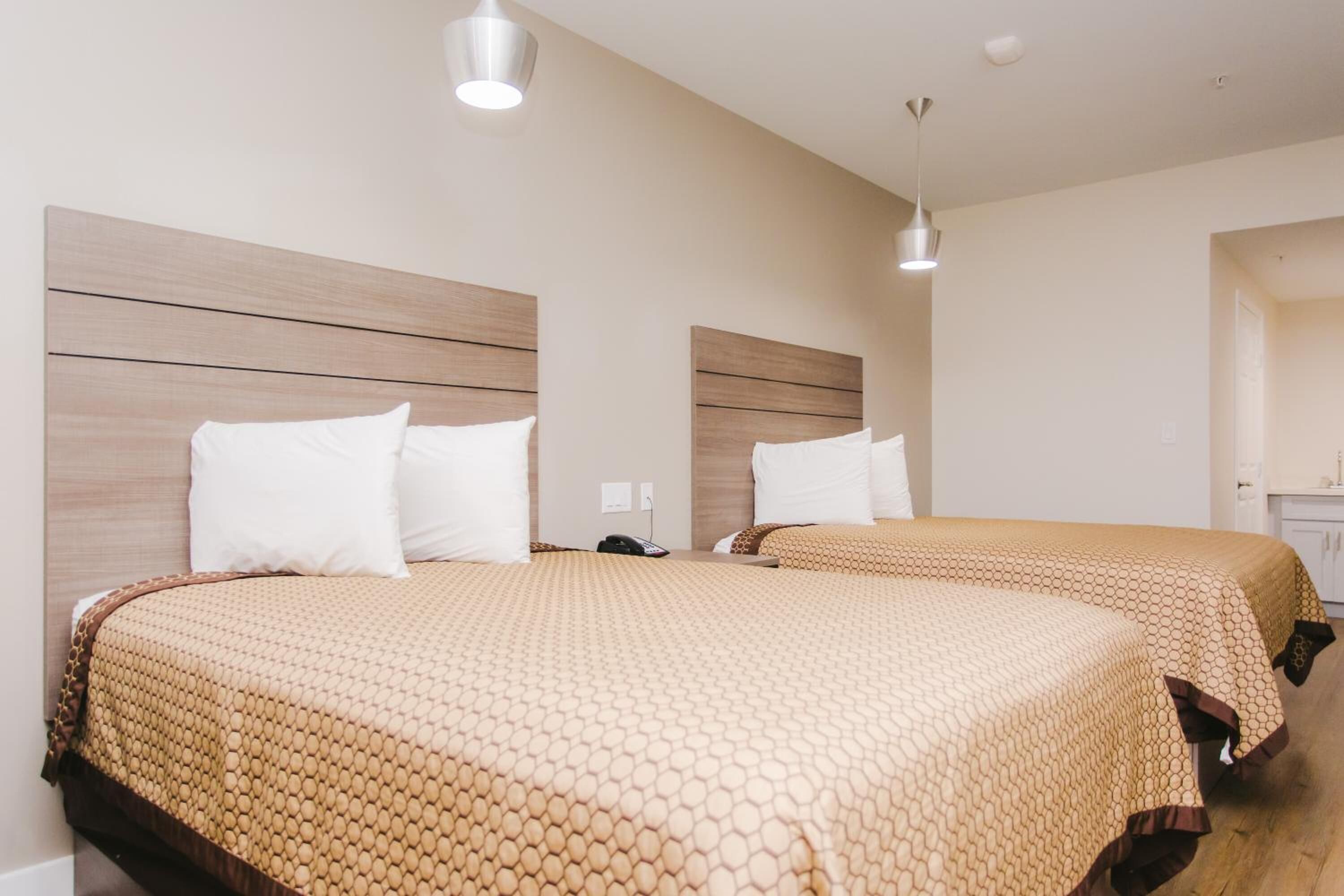 Una habitación de hotel con dos camas, cada una con cabecero de madera, colcha de color beige estampada y almohadas blancas. Dos lámparas colgantes están sobre las camas.