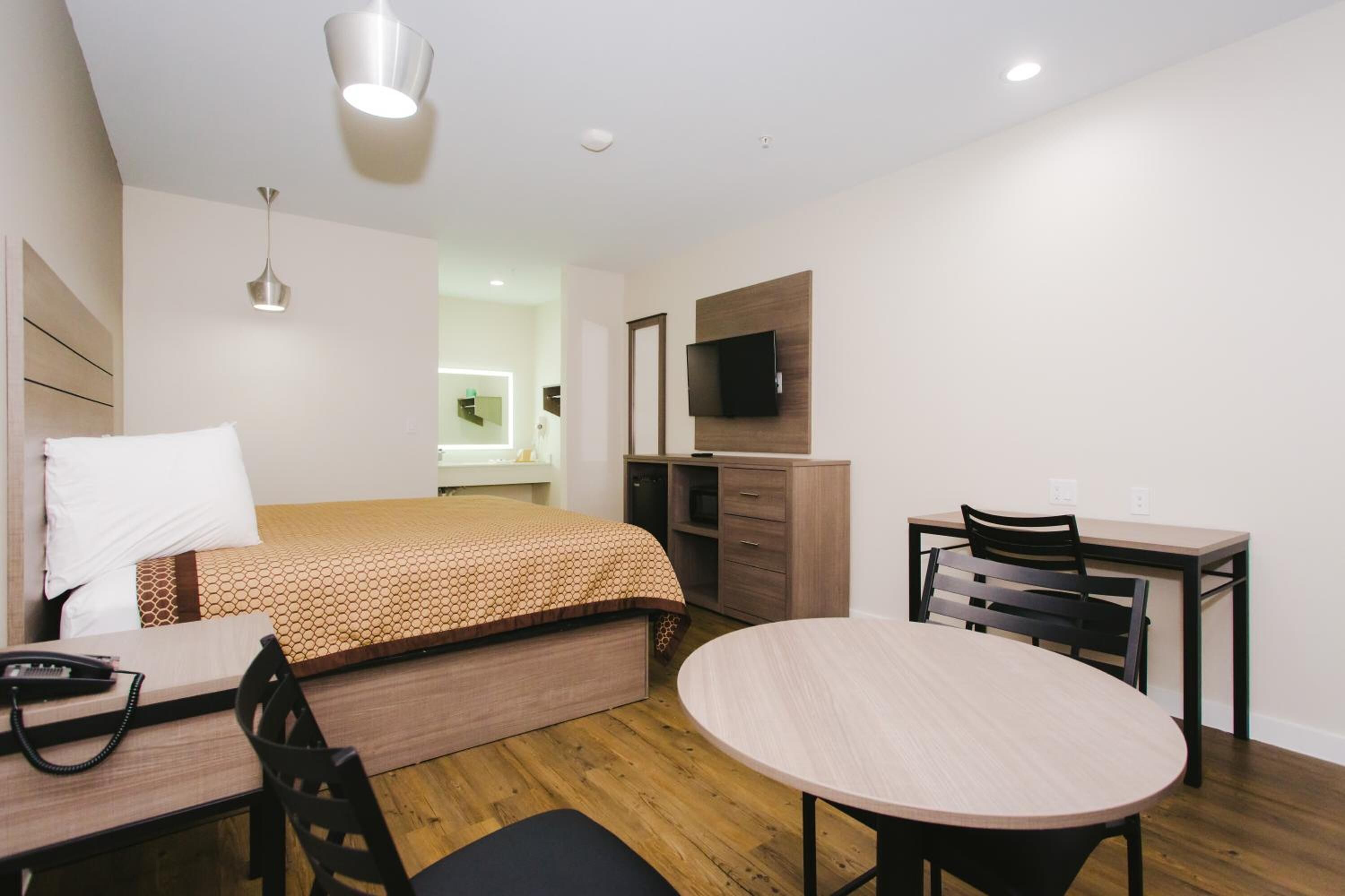 A modern hotel room with a bed, a desk with a chair, a round table, a TV mounted on the wall, and a hanging light fixture.