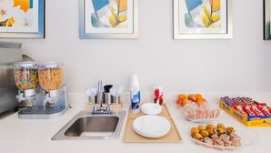 A kitchen counter with a sink, a coffee machine, a plate rack, and various food items including cookies and a box of Snickers. - Palace Inn - Louetta (Spring)
