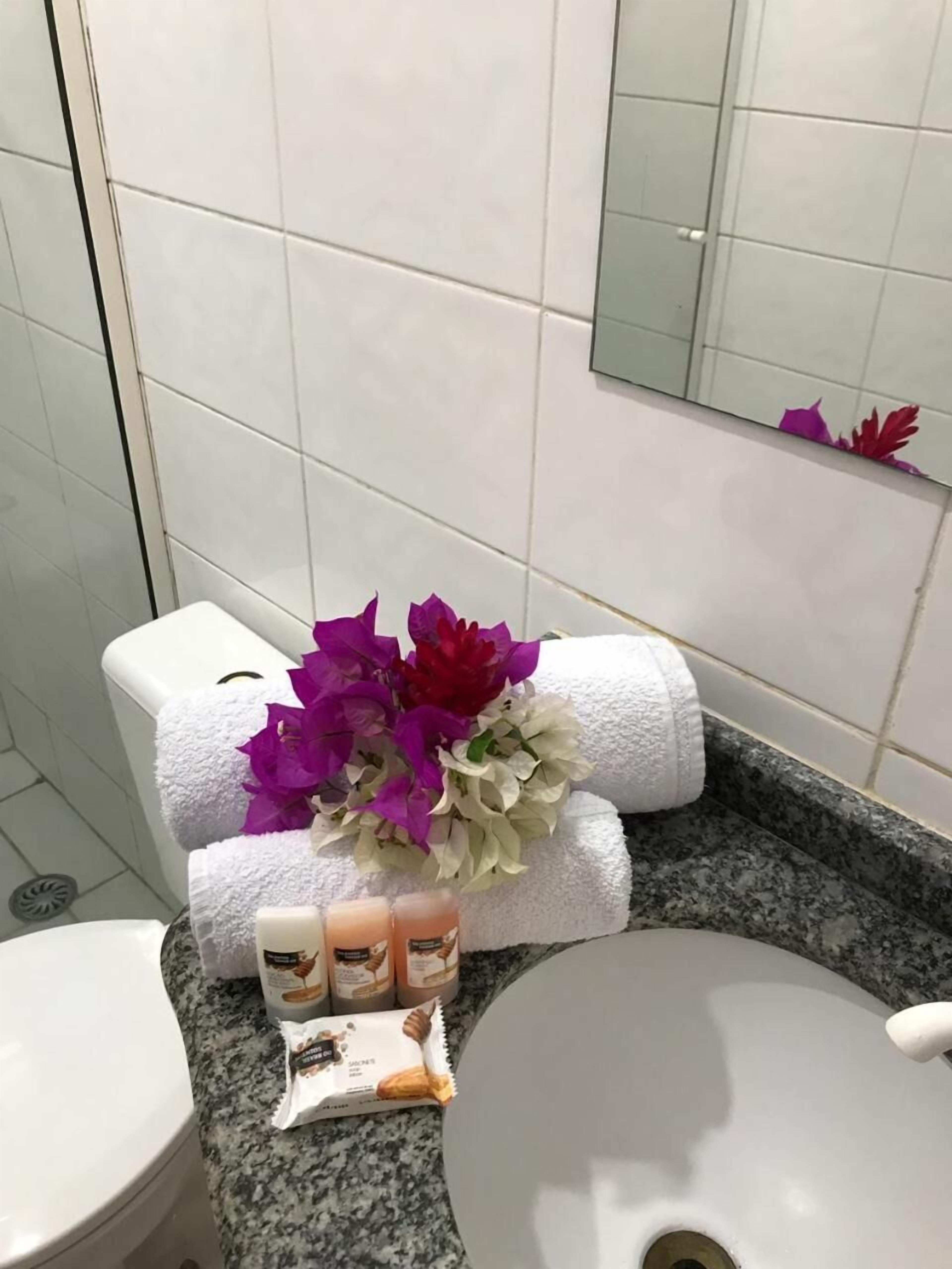 A bathroom with white tiles, a granite countertop, a white sink, a toilet with a white towel, and a bouquet of purple and white flowers.