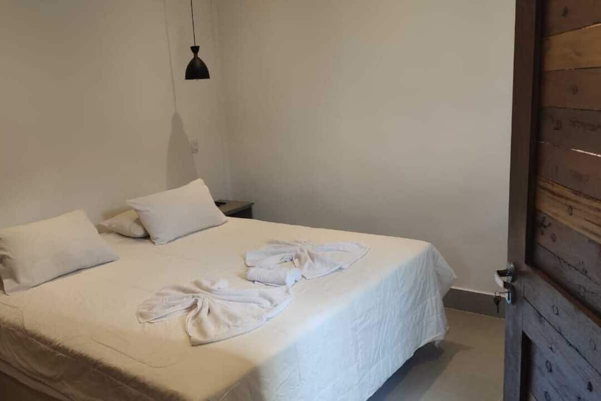 Een net opgemaakt bed met witte lakens en handdoeken, een zwart hanglamp en een houten deurkozijn.