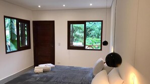 Une chambre avec un grand lit, deux fenêtres donnant sur un paysage verdoyant, un parquet foncé et un luminaire suspendu noir.